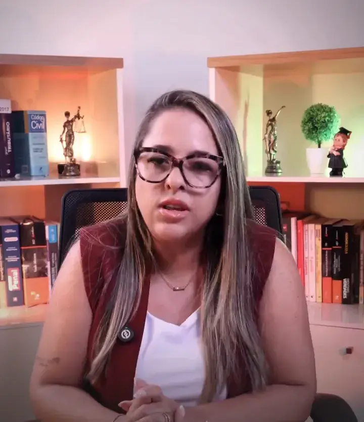 Dra. Keitty Machado, de óculos, colete vinho e blusa branca, explicando com um gesto das mãos, em frente a uma estante.